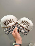 Two decorative hats with 'MAMA' and 'MINI' text, one leopard print, held by a hand against a neutral background.