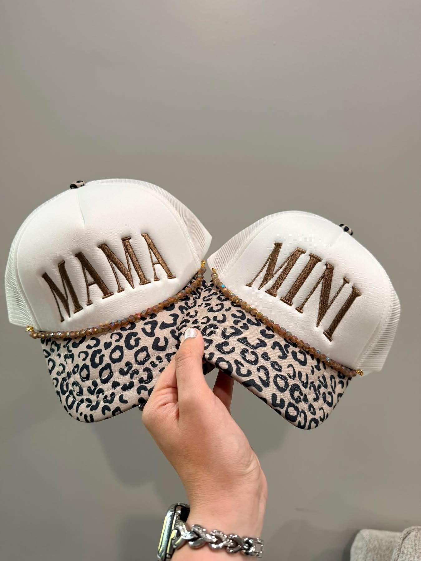 Two decorative hats with 'MAMA' and 'MINI' text, one leopard print, held by a hand against a neutral background.